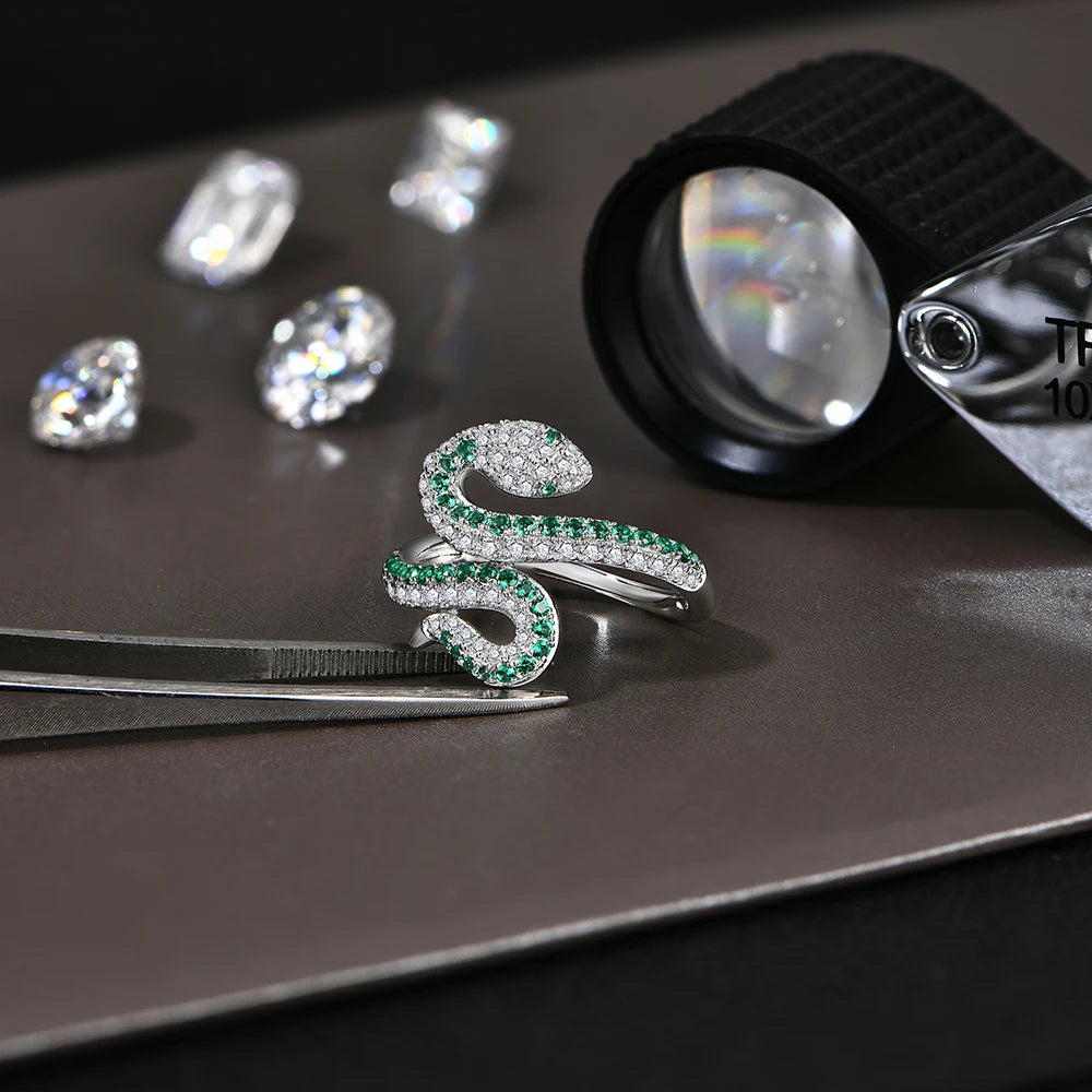 Silver snake-shaped ring with green gemstones on a dark surface with diamonds and a magnifying glass.