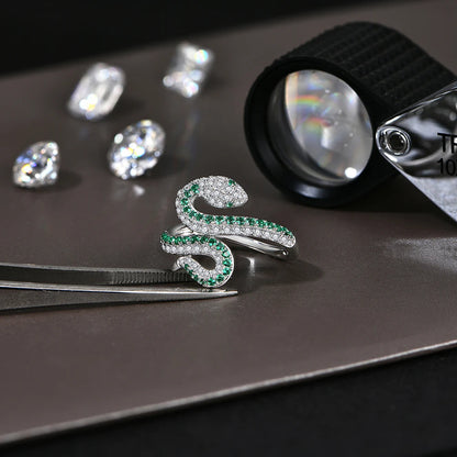 Silver snake-shaped ring with green gemstones on a dark surface with diamonds and a magnifying glass.