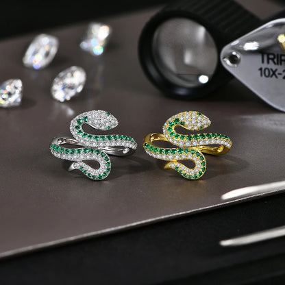 Two snake-shaped rings, one gold and one silver, with green gemstones on a reflective surface.